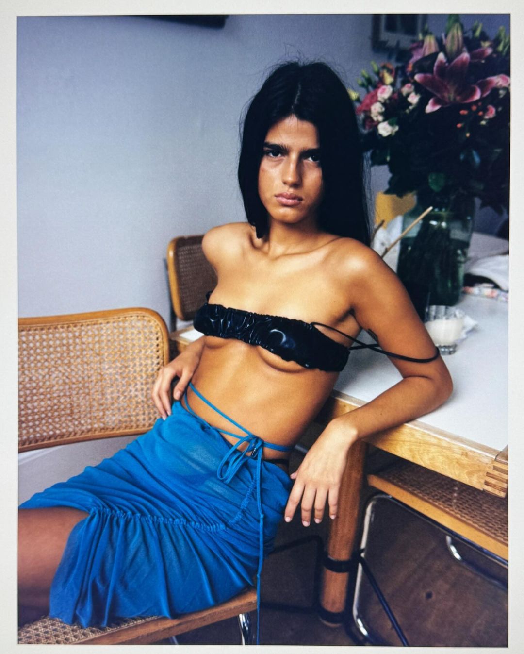 Woman in a black bandeau top and blue skirt seated at a table indoors by Angelo Pennetta
