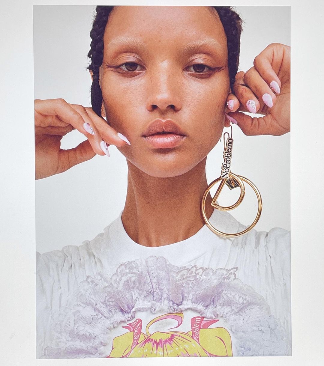 Close-up portrait of a woman with braided hair wearing large gold hoop earrings and light-colored nail polish against a white background.