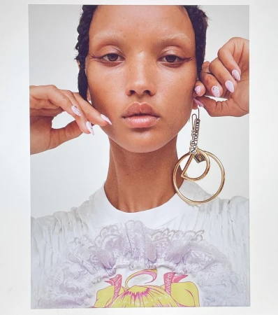 Close-up portrait of a woman with braided hair wearing large gold hoop earrings and light-colored nail polish against a white background.