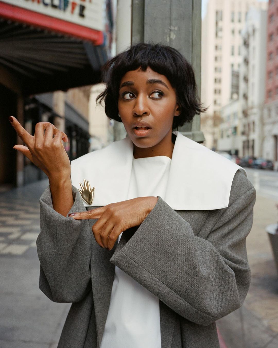 A young African-American woman stands on a city street in a gray coat, points with her left hand, and looks left with a surprised expression.
