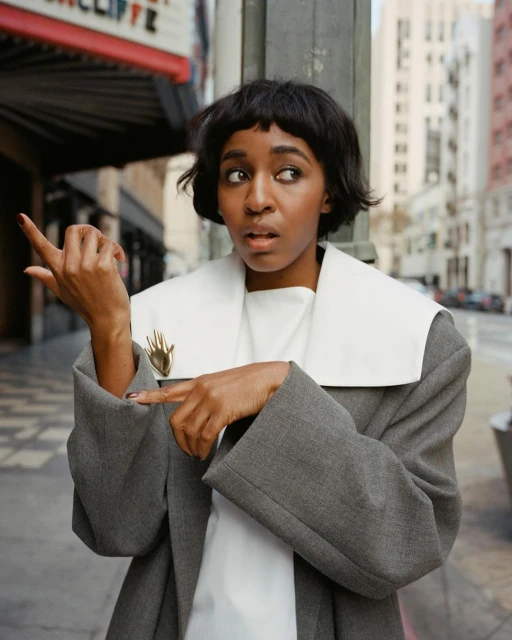 A young African-American woman stands on a city street in a gray coat, points with her left hand, and looks left with a surprised expression.
