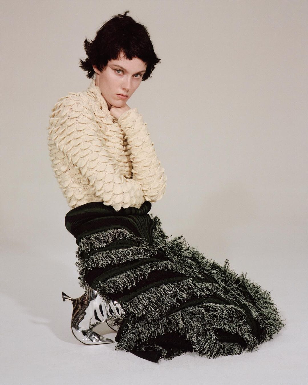 Fashion photo of a young adult with short dark hair, wearing a textured beige top, a black fringe skirt, and silver high heels, kneeling.