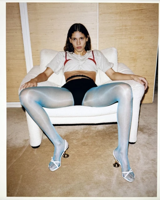 A woman sits in a chair wearing a gray t-shirt, black underwear, shiny blue tights, and white high heels in an indoor setting.