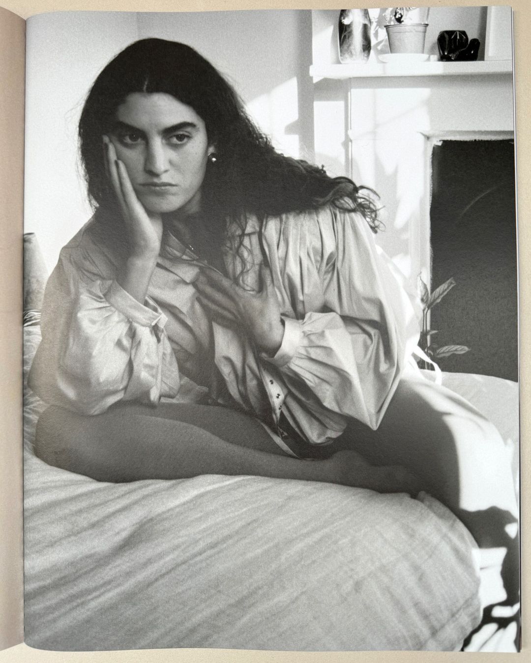 Black and white portrait of a woman with long dark hair seated on a bed, resting her head on her hand with a pensive expression.