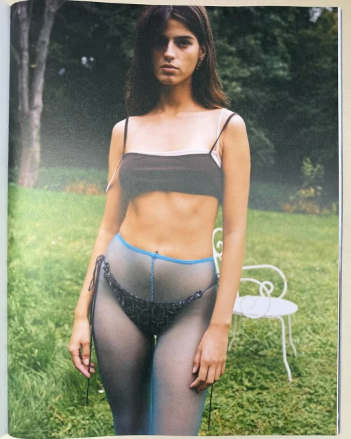 A young woman poses outdoors wearing a brown crop top and sheer pantyhose, standing in a natural setting with greenery and a white chair.