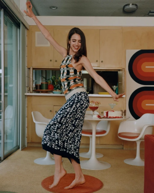 A young woman in patterned clothing dances barefoot in a sunlit interior, with a table, cocktails, and retro decor visible.