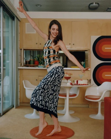 A young woman in patterned clothing dances barefoot in a sunlit interior, with a table, cocktails, and retro decor visible.