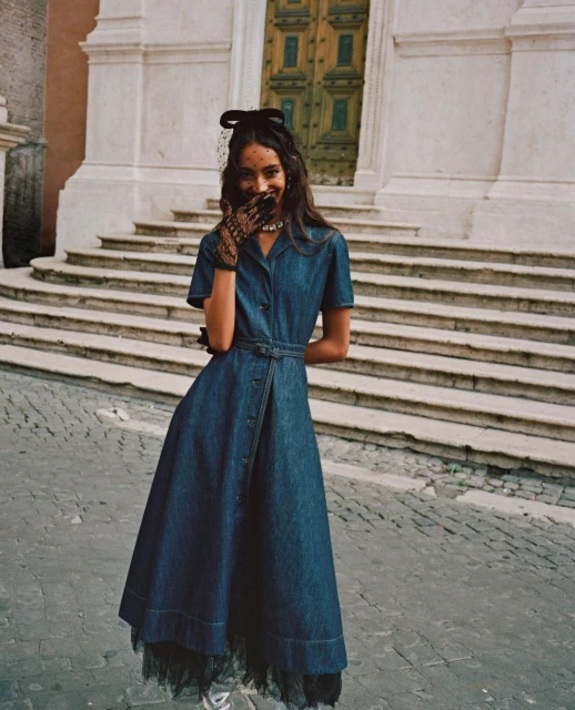 A woman in a denim dress, wearing a netted headpiece and lace gloves, smiles in front of stone steps leading to an arched doorway.