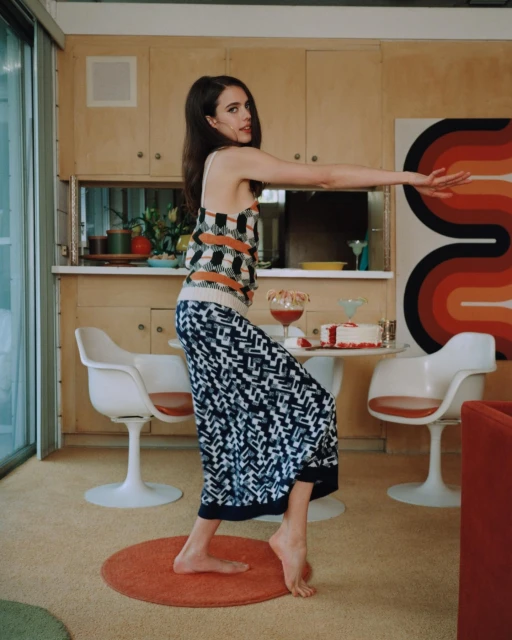 A woman with long dark hair poses in a patterned outfit, indoors, against a retro-style kitchen backdrop with a white tulip table.