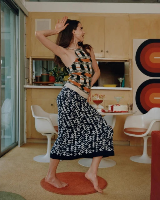 A barefoot woman dances in a stylish kitchen wearing a patterned top and skirt, with mid-century modern decor and soft, warm lighting.