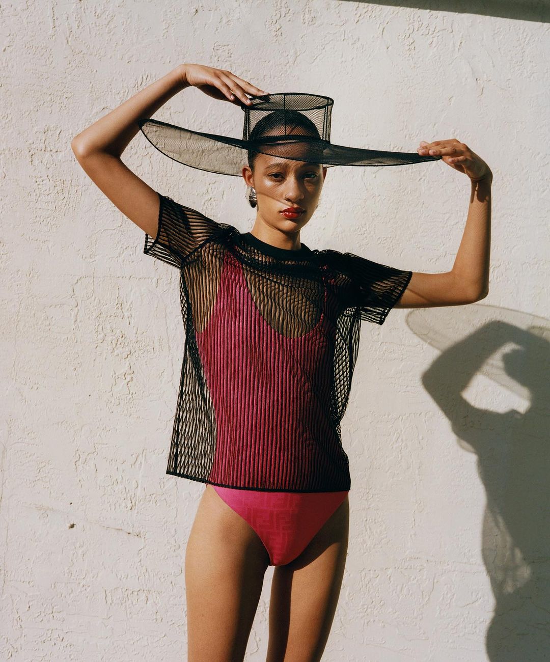 A young woman in a pink swimsuit and black mesh top poses against a white wall wearing a large mesh hat, fashion shot.