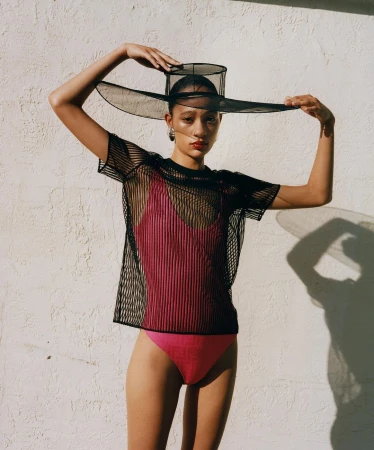 A young woman in a pink swimsuit and black mesh top poses against a white wall wearing a large mesh hat, fashion shot.