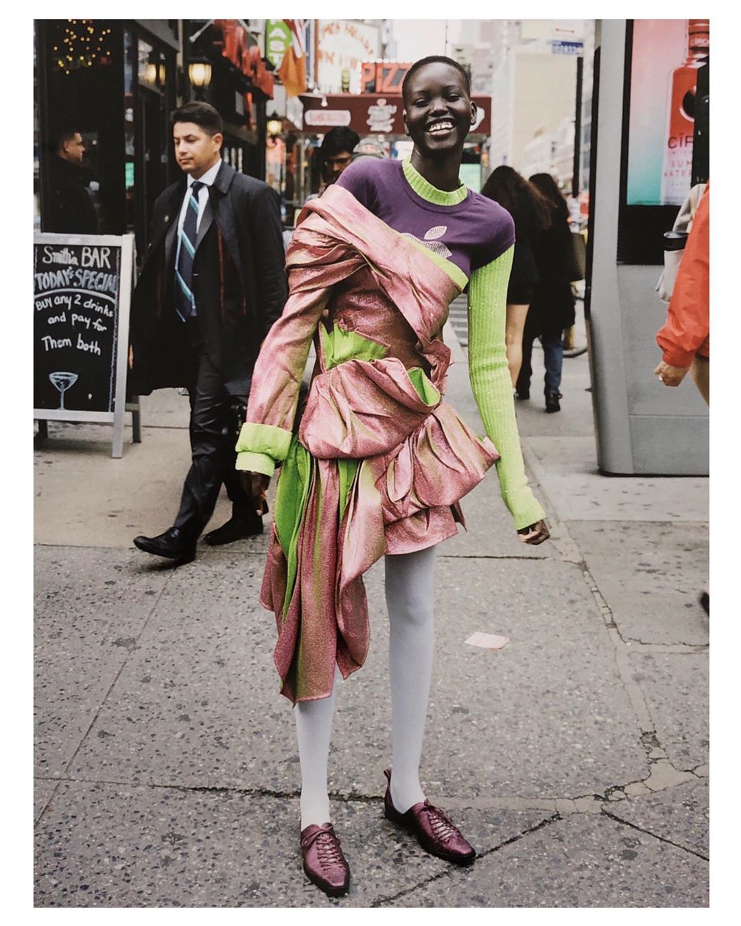 Smiling African woman wears a pink and green dress on a city street with pedestrians and a sandwich board in the background.