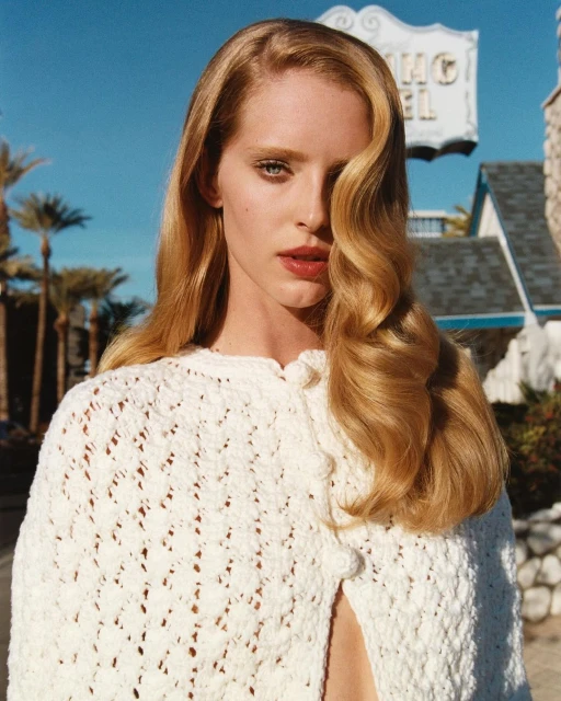 Portrait of a blonde woman with long wavy hair wearing a white crochet poncho in front of the El Rey Hotel and palm trees.