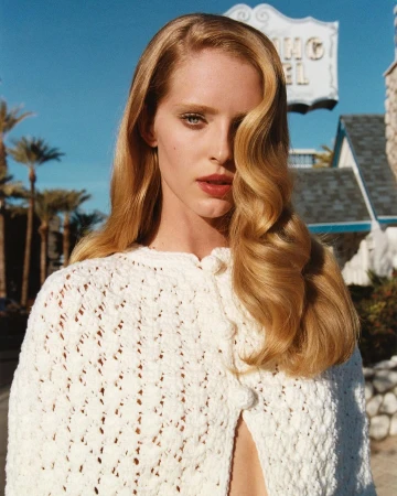 Portrait of a blonde woman with long wavy hair wearing a white crochet poncho in front of the El Rey Hotel and palm trees.