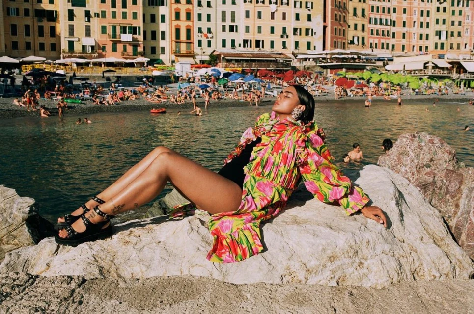 A woman reclines on a rock near the shore in a ruffled floral dress and sandals. The beach is filled with people and buildings in the distance.