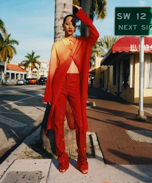 Fashionable adult woman poses in a two-tone top, red pants and heels on a city sidewalk with palm trees and buildings in the background.