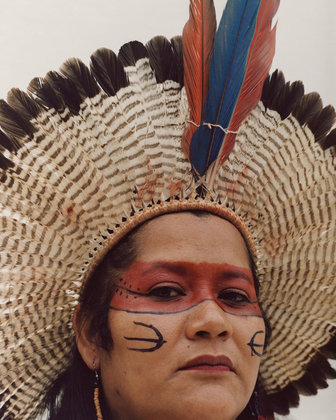 Close-up of an indigenous woman with face paint and a large feathered headdress against a neutral background in a portrait style.