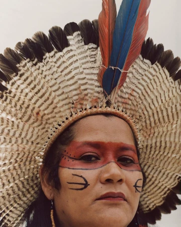 Close-up of an indigenous woman with face paint and a large feathered headdress against a neutral background in a portrait style.