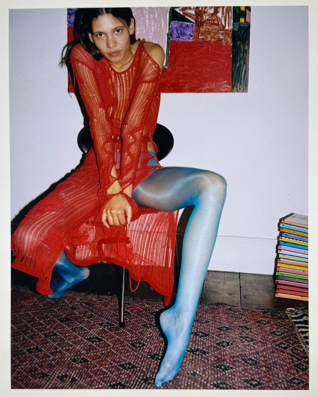 A woman in a red knit dress sits on a chair in a studio setting, with a painting and books in the background and blue tights.