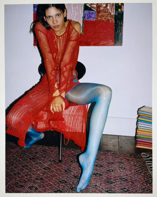 A woman in a red knit dress sits on a chair in a studio setting, with a painting and books in the background and blue tights.