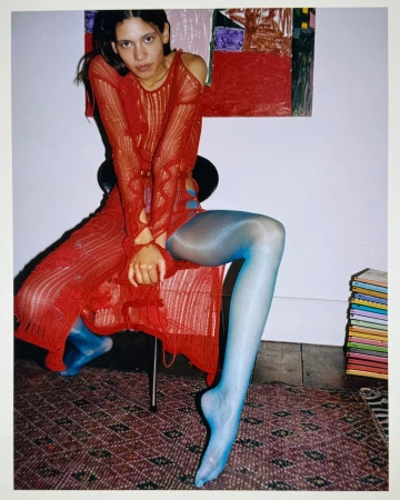 A woman in a red knit dress sits on a chair in a studio setting, with a painting and books in the background and blue tights.