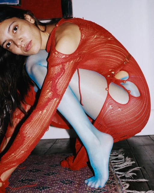 Woman in a red, open-knit dress poses with blue stockings on a patterned rug against a white wall in a studio setting.