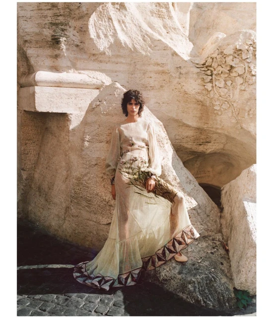 Fashion shot of a woman in a dress, posing in front of a travertine stone structure, creating an elegant and timeless scene.
