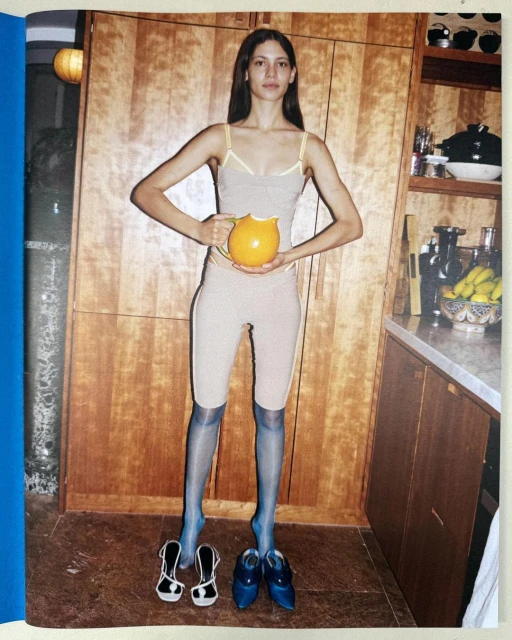 Woman in beige top and leggings stands in a kitchen holding an orange pitcher, with several pairs of shoes on the floor.