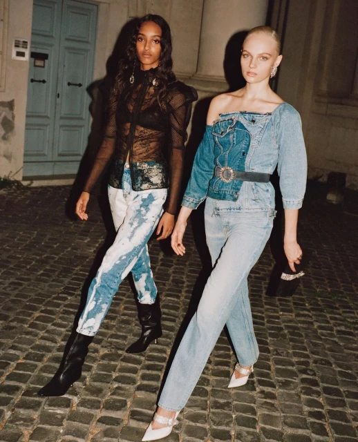 Two fashion models in stylish denim and lace ensembles pose outdoors on a cobblestone street, illuminated by soft evening light.