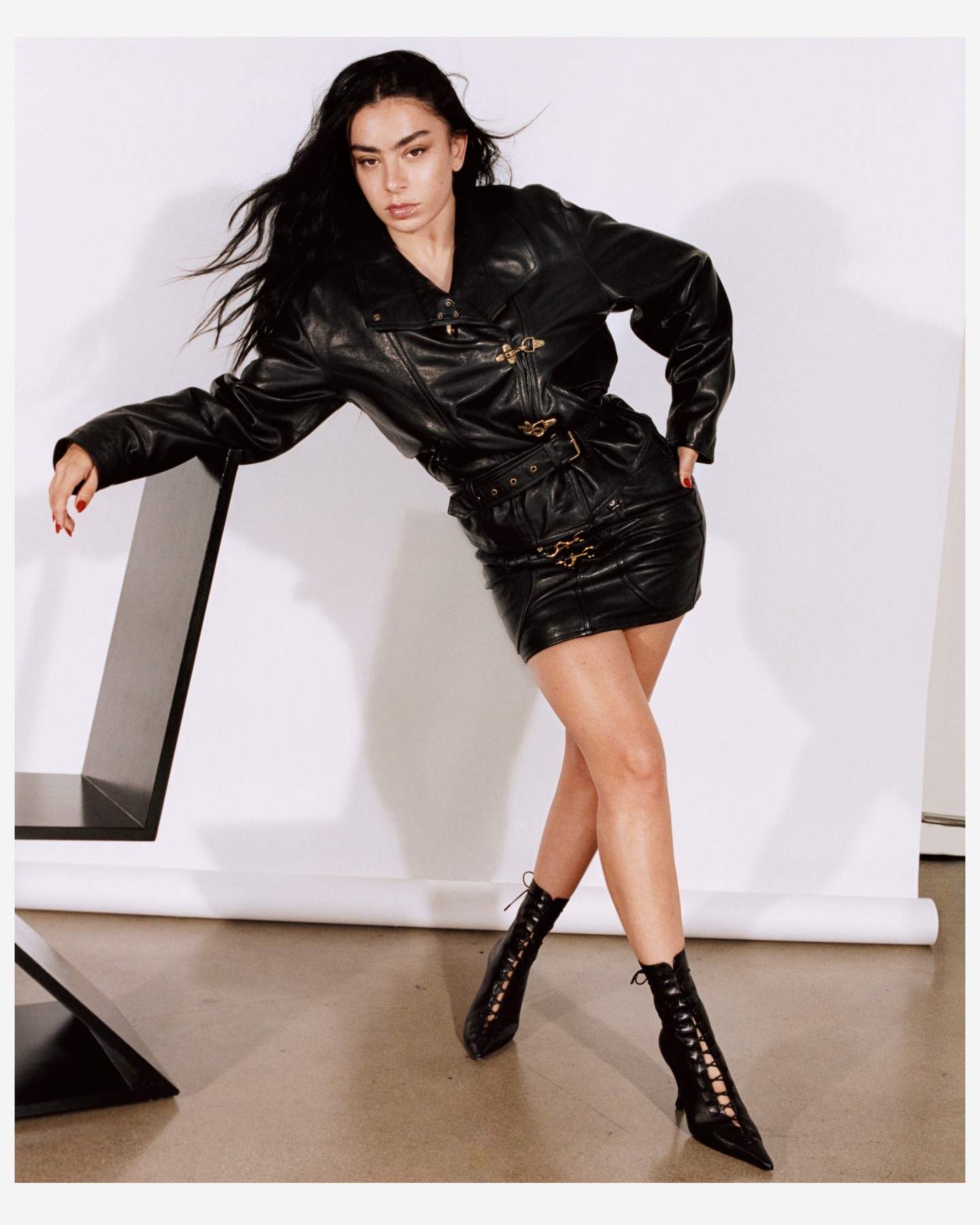 A fashionable woman with long dark hair poses in a black leather jacket and matching mini skirt in a studio setting.
