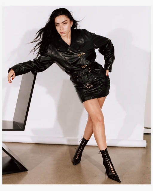 A fashionable woman with long dark hair poses in a black leather jacket and matching mini skirt in a studio setting.