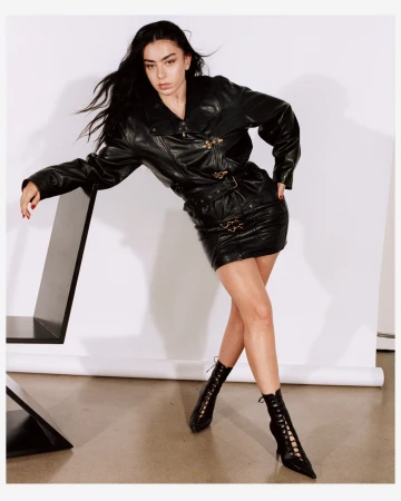 A fashionable woman with long dark hair poses in a black leather jacket and matching mini skirt in a studio setting.