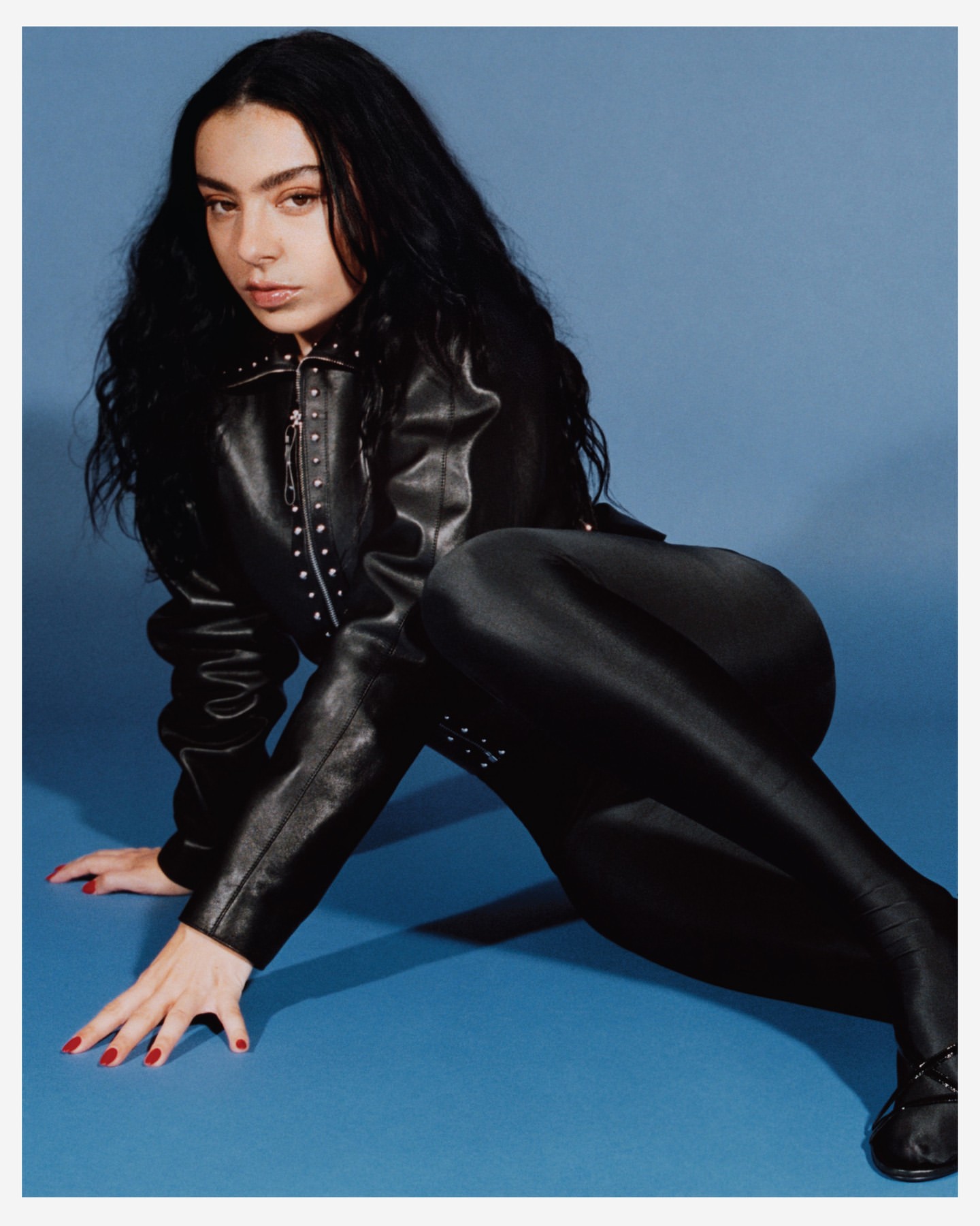 A woman with long dark hair poses on a blue background, wearing a studded leather jacket and matching shiny pants and boots.