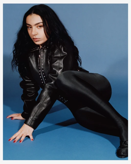 A woman with long dark hair poses on a blue background, wearing a studded leather jacket and matching shiny pants and boots.