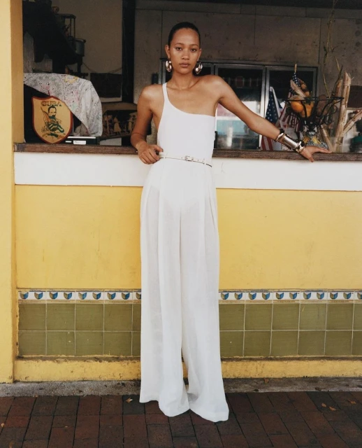 A woman poses in an all-white one-shoulder jumpsuit with gold hoop earrings, a matching bracelet, and a slim belt outdoors against a yellow wall.
