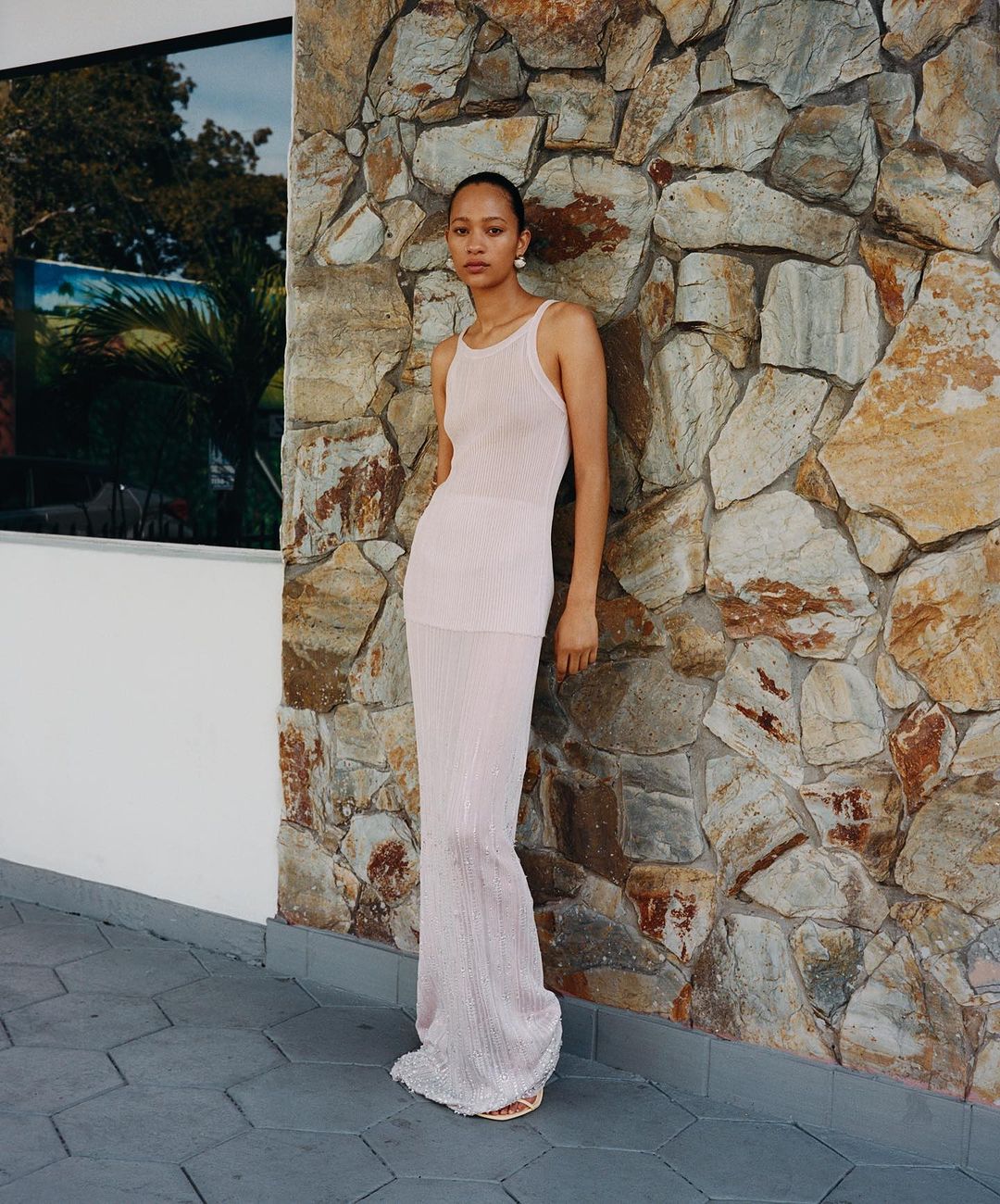 Young woman in a light pink knit dress stands against a textured stone wall. Tropical foliage is reflected in a window nearby.