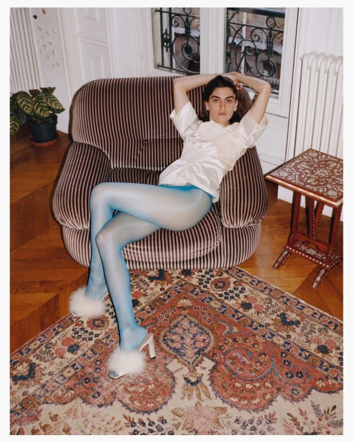 Adult female fashion model poses in a satin blouse, tights, and fuzzy mules on a striped chair over an ornate rug inside an apartment.