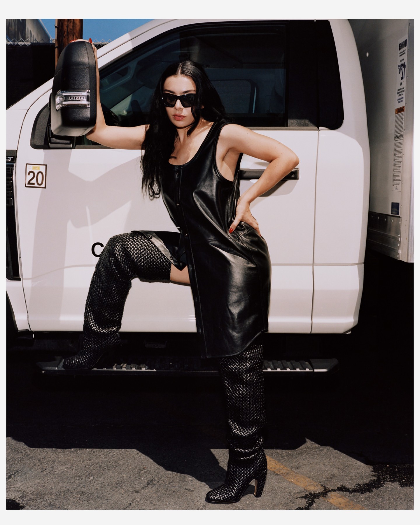 Woman in black leather dress and sunglasses posing against a white truck