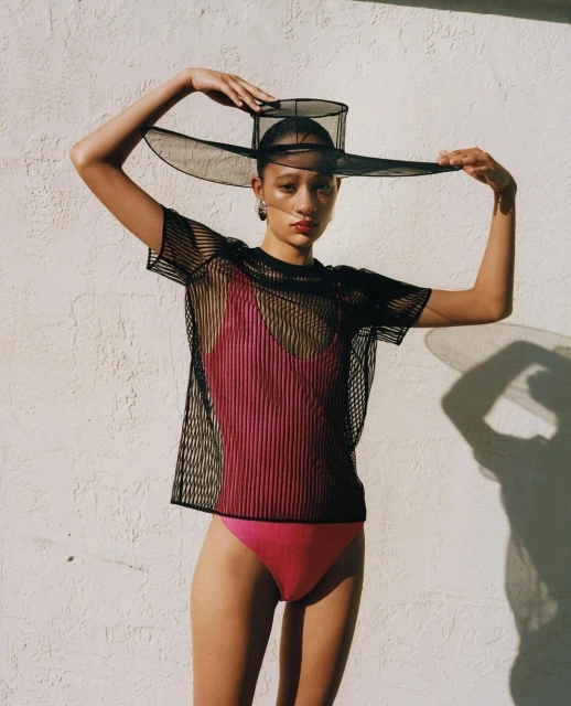 Woman wearing a mesh top and a wide-brimmed hat poses against a textured off-white wall, casting shadows in an outdoor setting.