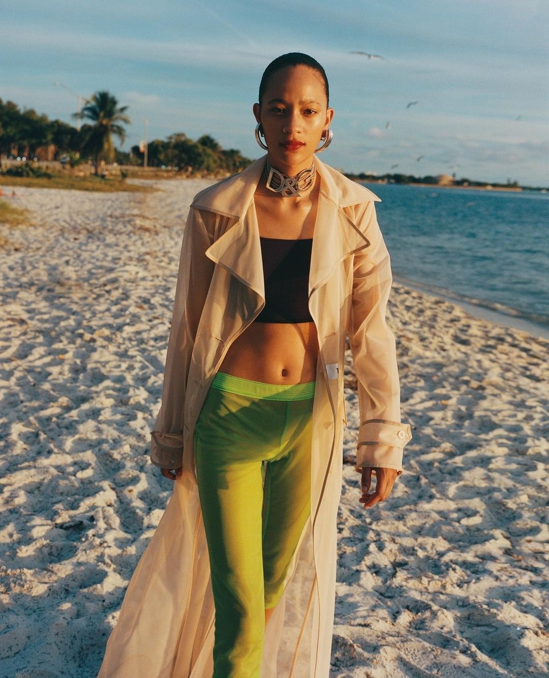 An adult female model walks on a sandy beach near the ocean, wearing a tan trench coat, black crop top, and lime green pants.
