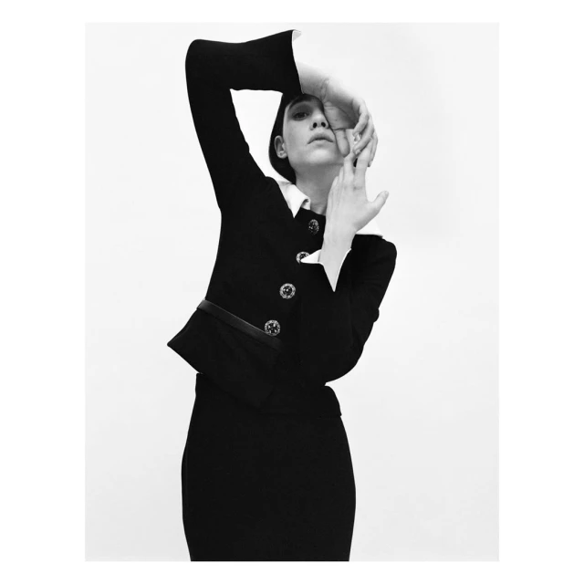 Monochrome portrait of a woman in a black suit jacket and skirt against a white background with her hands near her face.