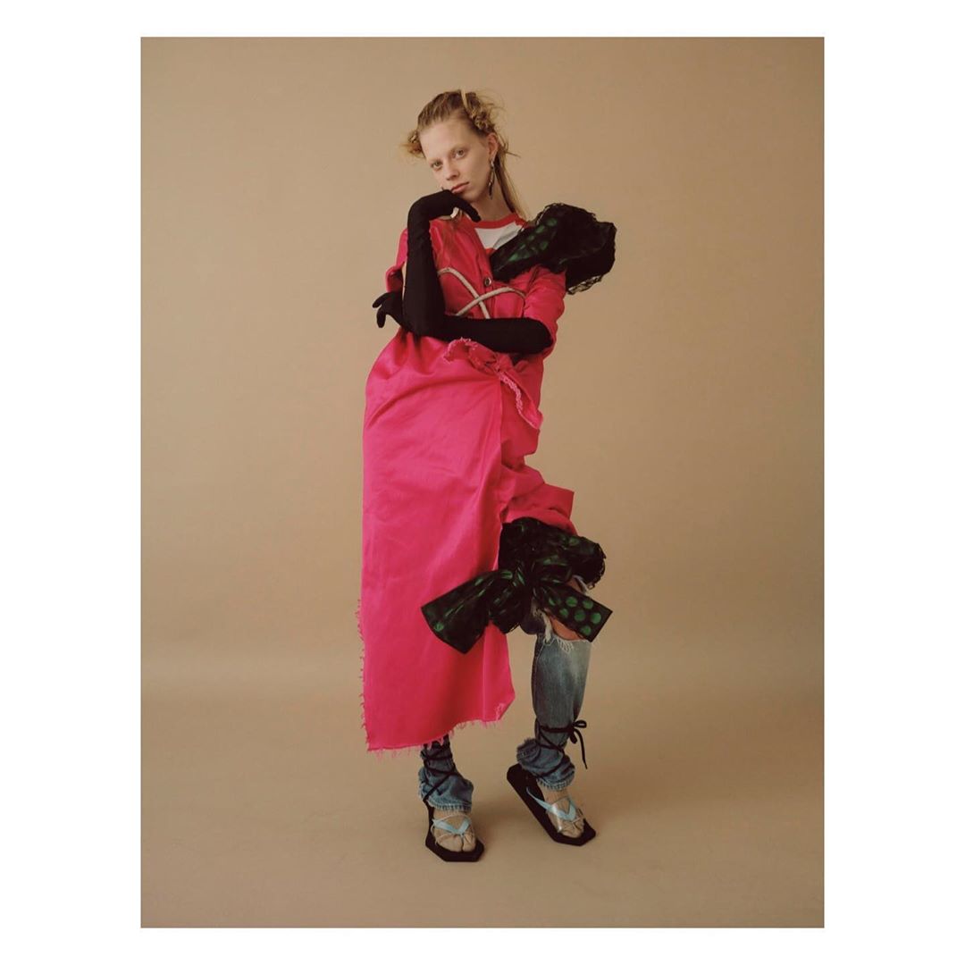 Fashion photo of a young, fair-skinned woman in a pink robe and denim jeans, posing against a neutral backdrop in a studio setting.