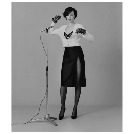 A woman in a black skirt and white sweater stands next to a microphone on a stand, wearing gloves, in a full studio shot against a gray backdrop.
