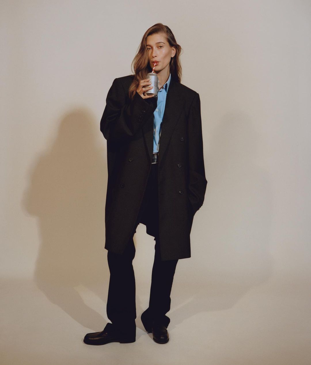 Adult female in a black oversized coat poses while drinking with a straw from a can against a neutral backdrop in a studio shot.