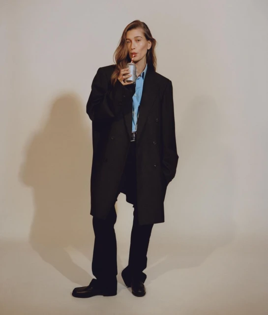 Adult female in a black oversized coat poses while drinking with a straw from a can against a neutral backdrop in a studio shot.