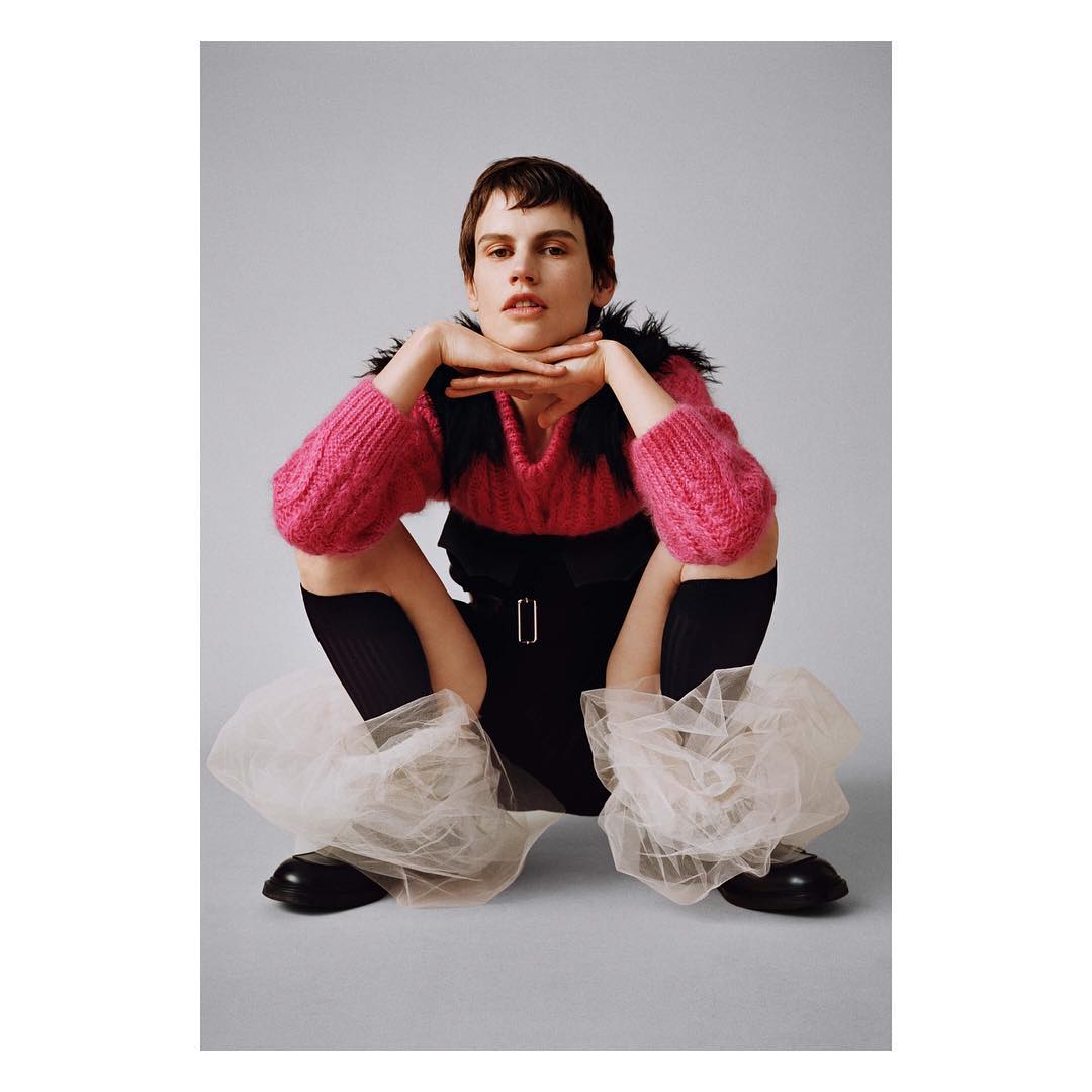 Model with short dark hair squatting in a pink sweater and black outfit with sheer tulle leg warmers