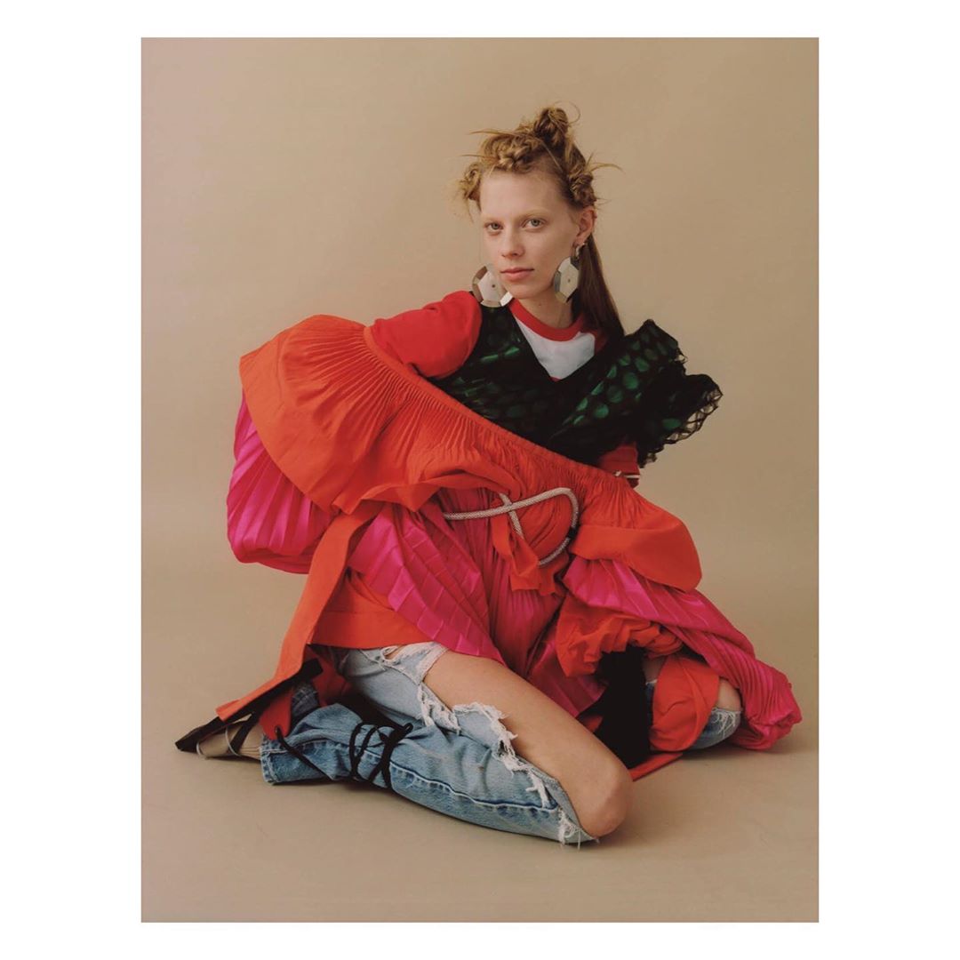 Stylish woman in a vibrant red and pink layered dress with ripped jeans, sitting in a studio with a neutral background, fashion editorial.