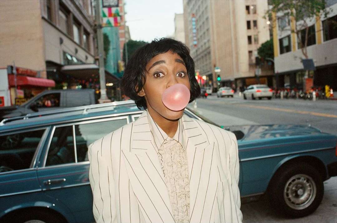 Woman in a striped blazer blowing a bubblegum bubble in an urban setting with a classic car, creating a vintage editorial style image.