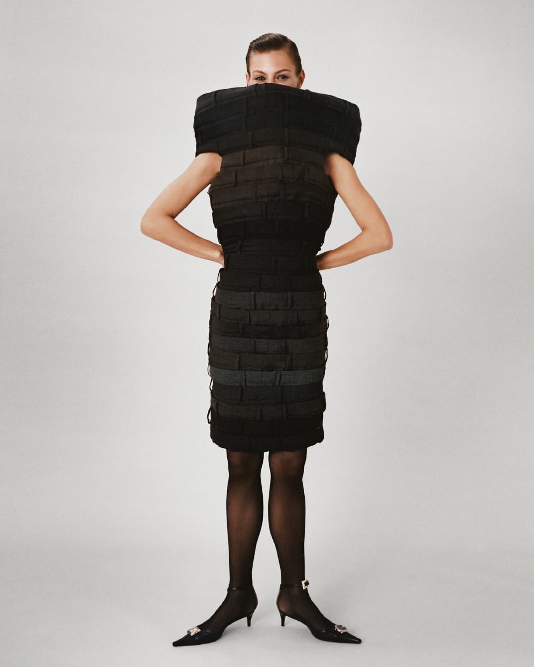 A woman poses in a structured black dress against a white backdrop, accessorized with black tights and pointed heels with jeweled embellishments.